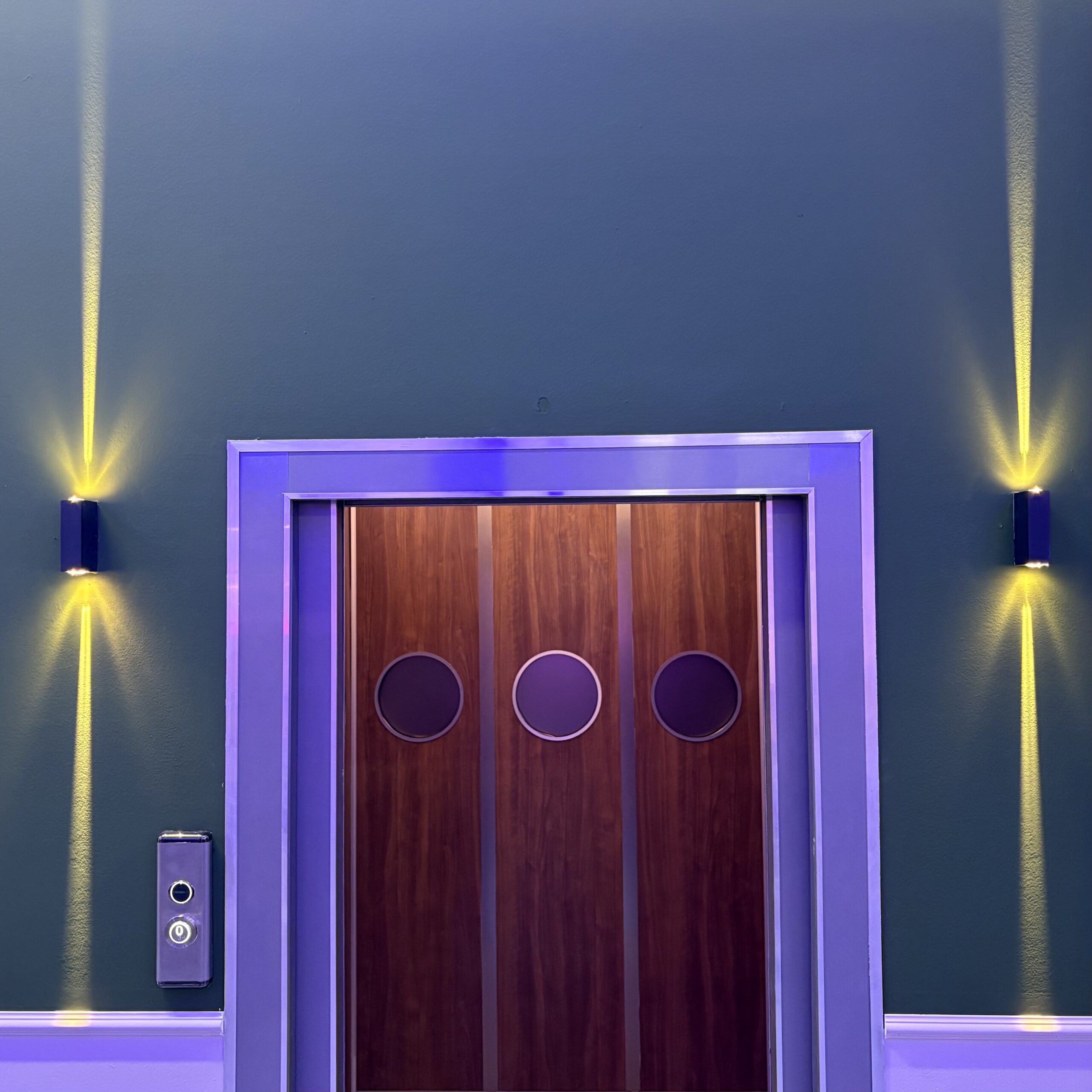 Porta ascensore scenografica in legno con tre oblò, cornice viola illuminata, pannello di controllo e applique a parete.