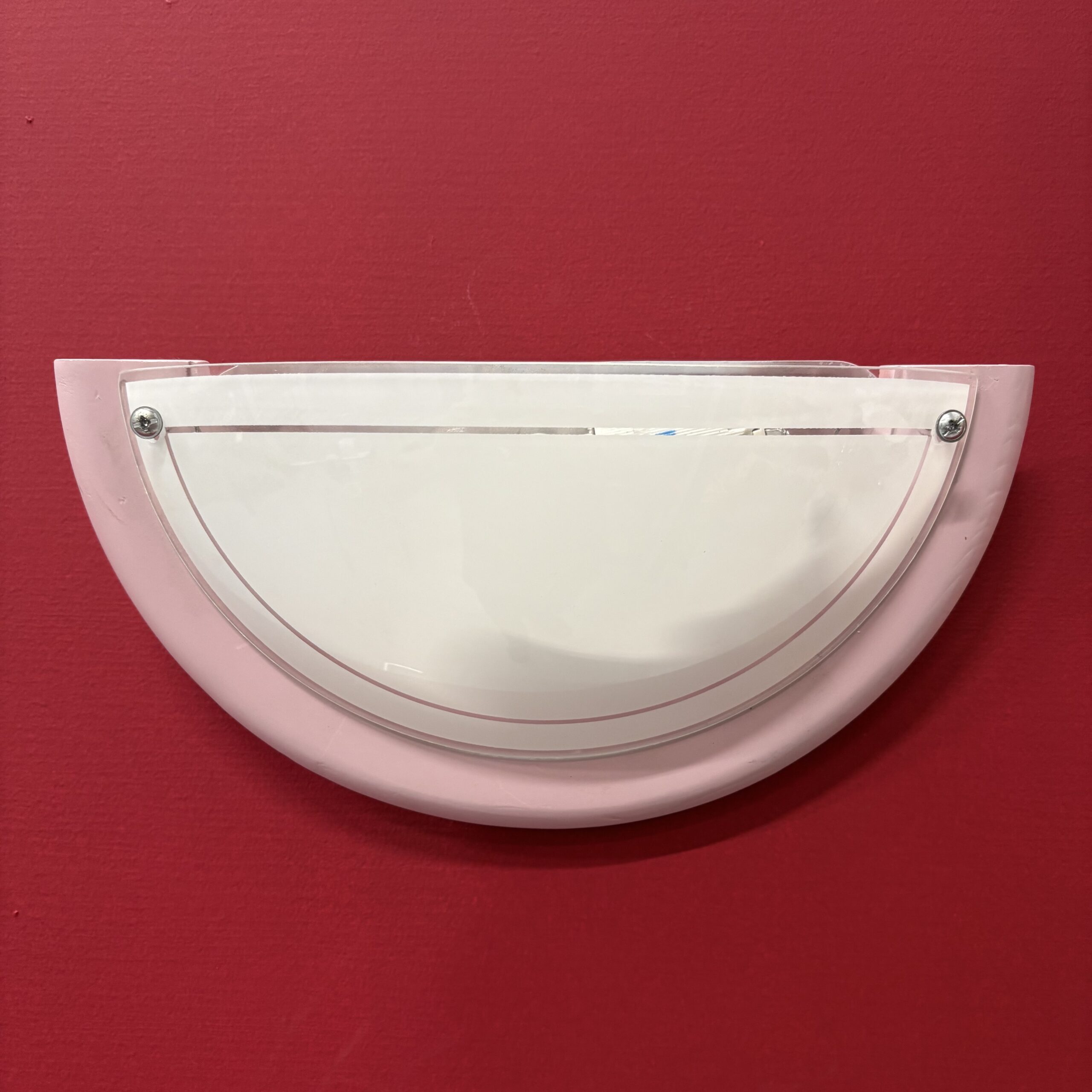 Applique da parete semilunare con base rosa pastello e diffusore in acrilico bianco traslucido, fissata su una parete rossa.