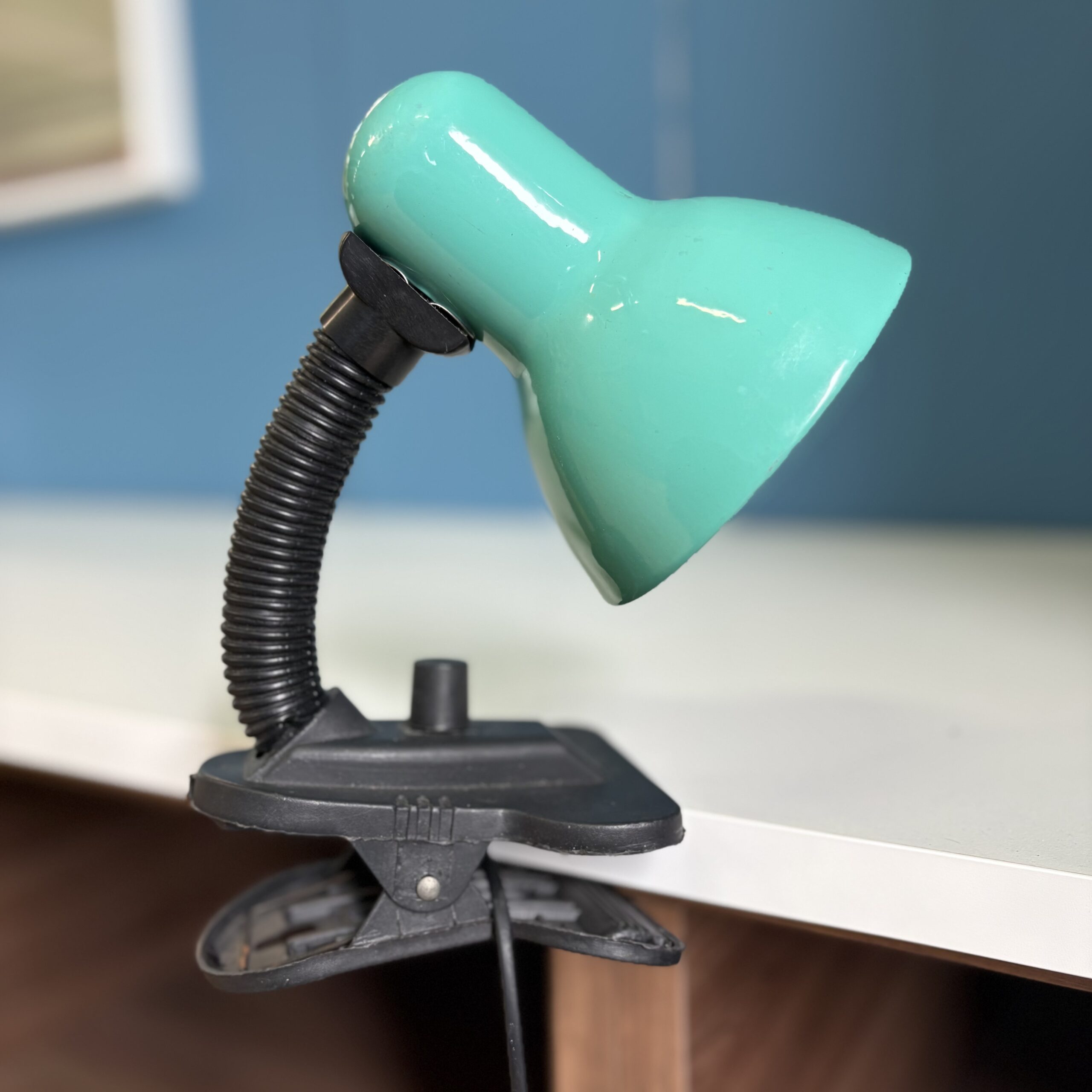 Lampada da tavolo a pinza con paralume verde acqua e braccio flessibile nero, fissata su un bordo bianco.