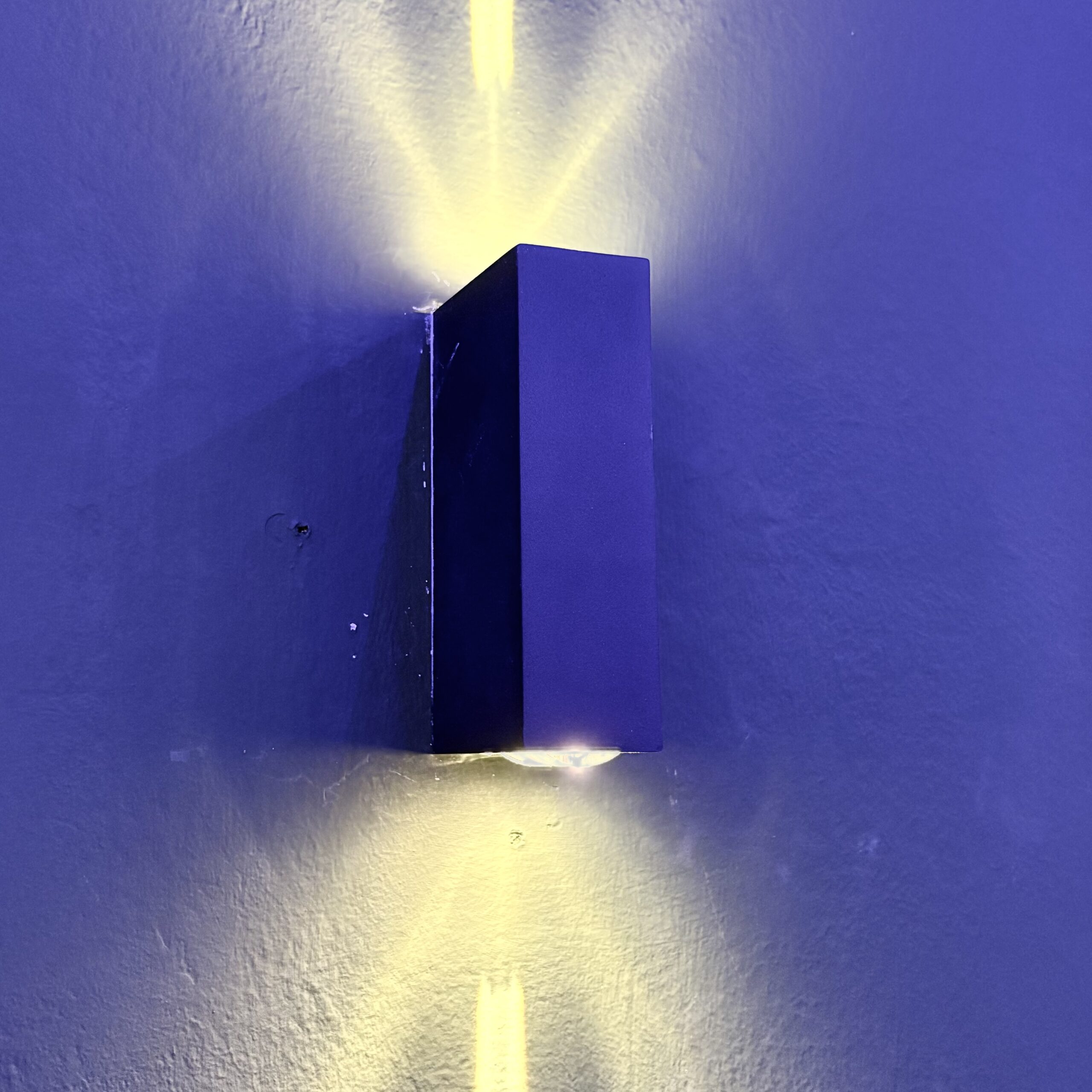 Applique LED bidirezionale nera montata su parete blu, che emette fasci di luce gialla verso l'alto e il basso.
