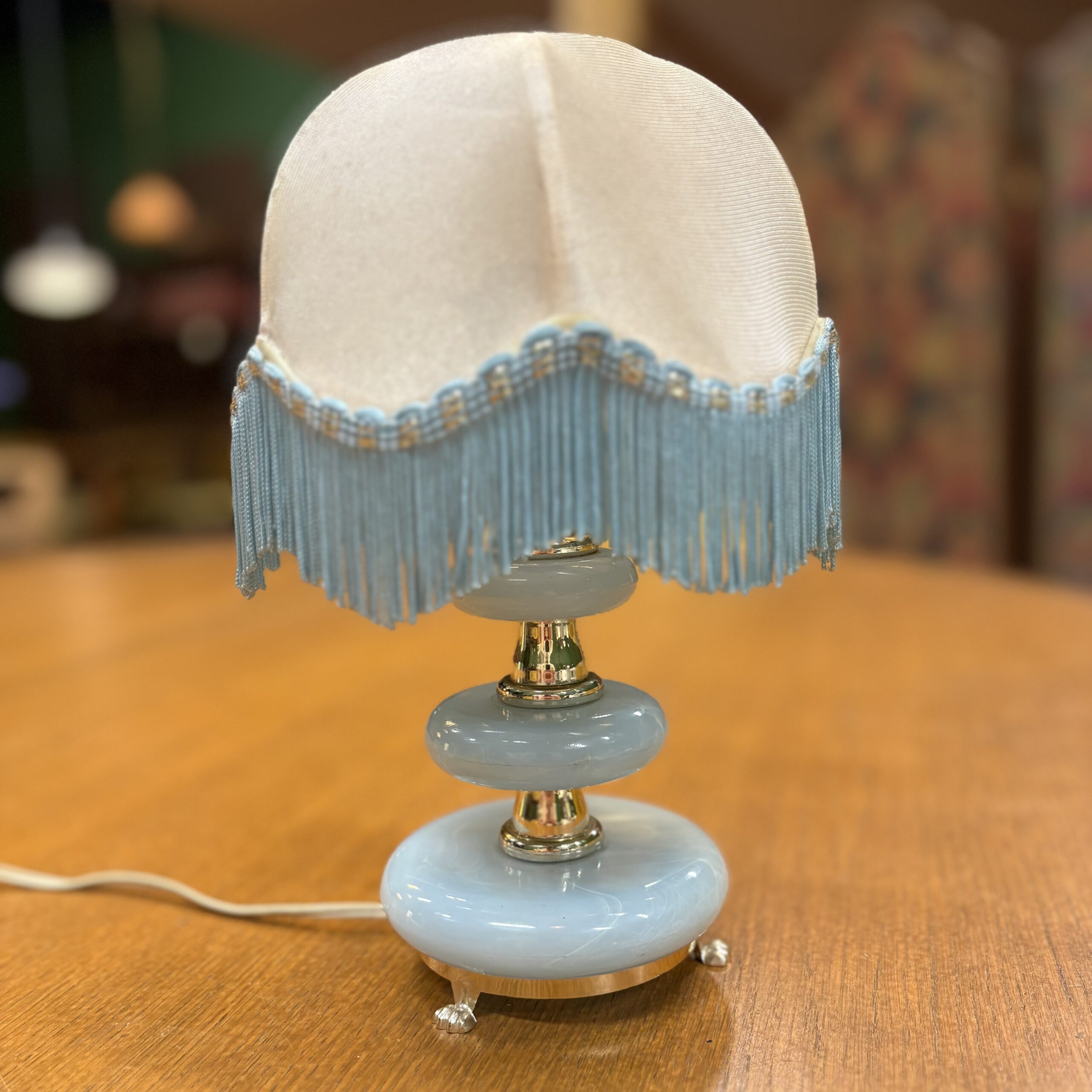 Lampada da tavolo vintage con base azzurra lucida, dettagli dorati e paralume in tessuto crema con frange azzurre.
