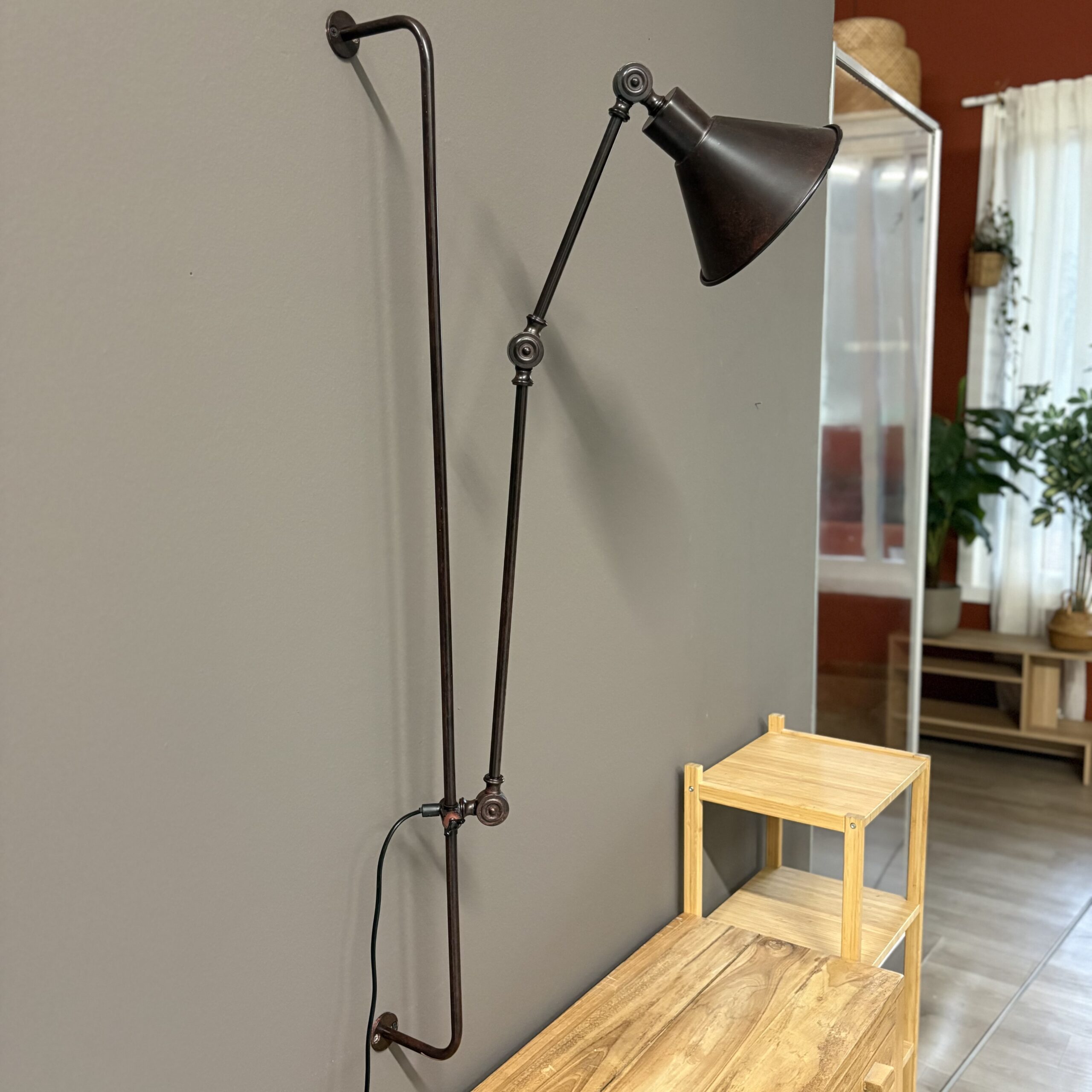 Lampada da parete industriale con braccio articolato e paralume a cono, finitura effetto bronzo invecchiato, montata su parete grigia.