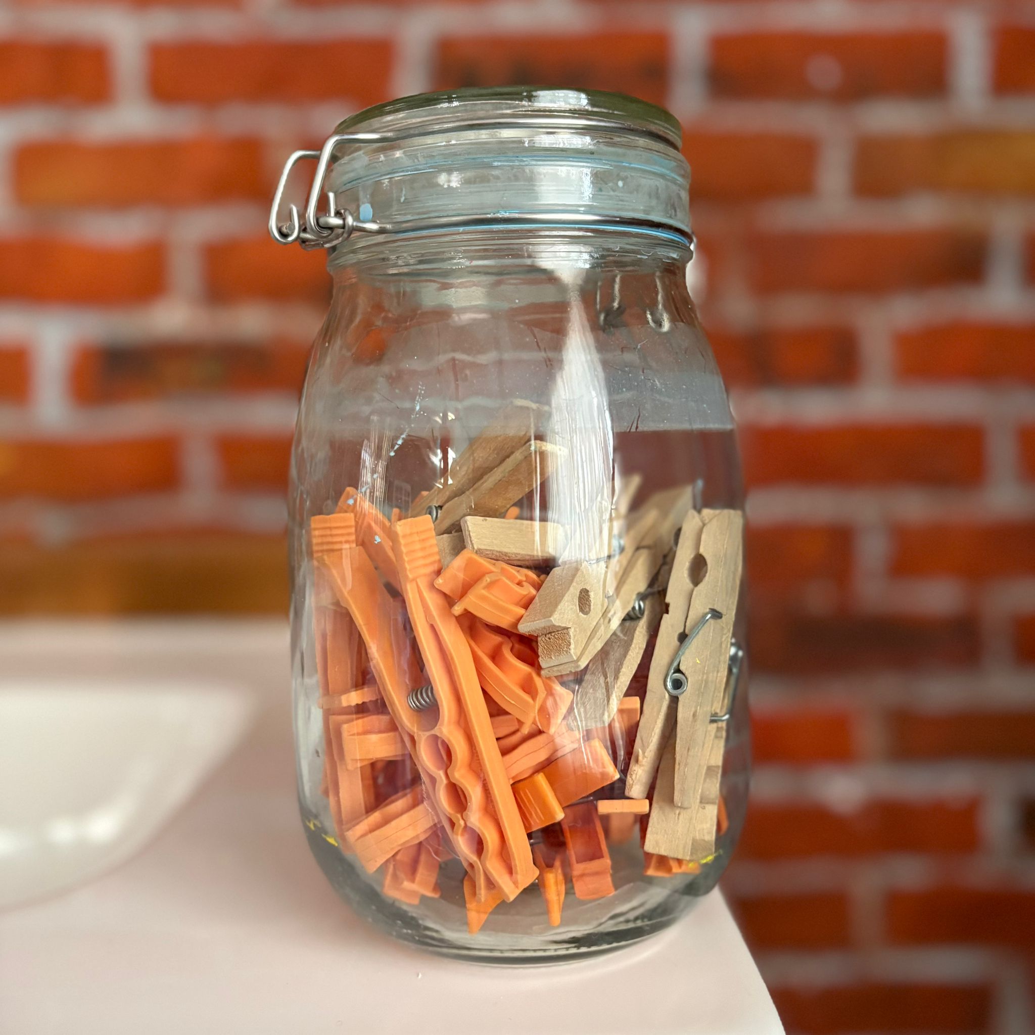 Barattolo in vetro trasparente con coperchio a chiusura ermetica, riempito con mollette da bucato in legno e plastica arancione, su sfondo di mattoni.