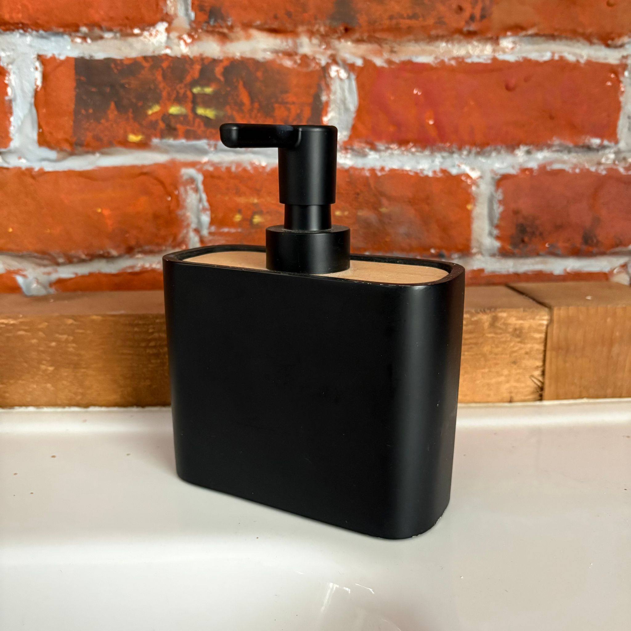 Dispenser per sapone liquido nero opaco con coperchio in legno chiaro e pompa nera, su superficie bianca con muro di mattoni.