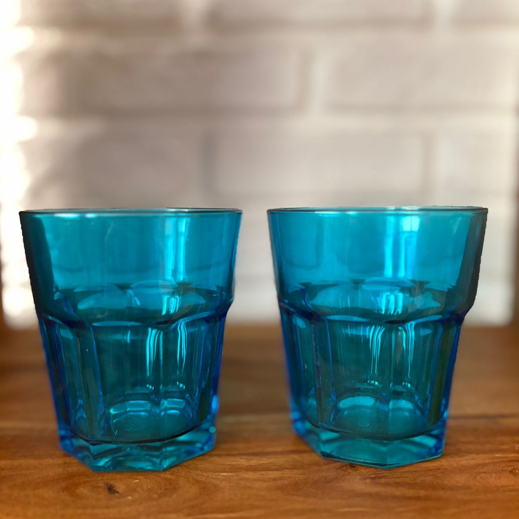 Due bicchieri tumbler in vetro blu sfaccettato, posizionati su una superficie di legno con uno sfondo di mattoni bianchi sfocati.