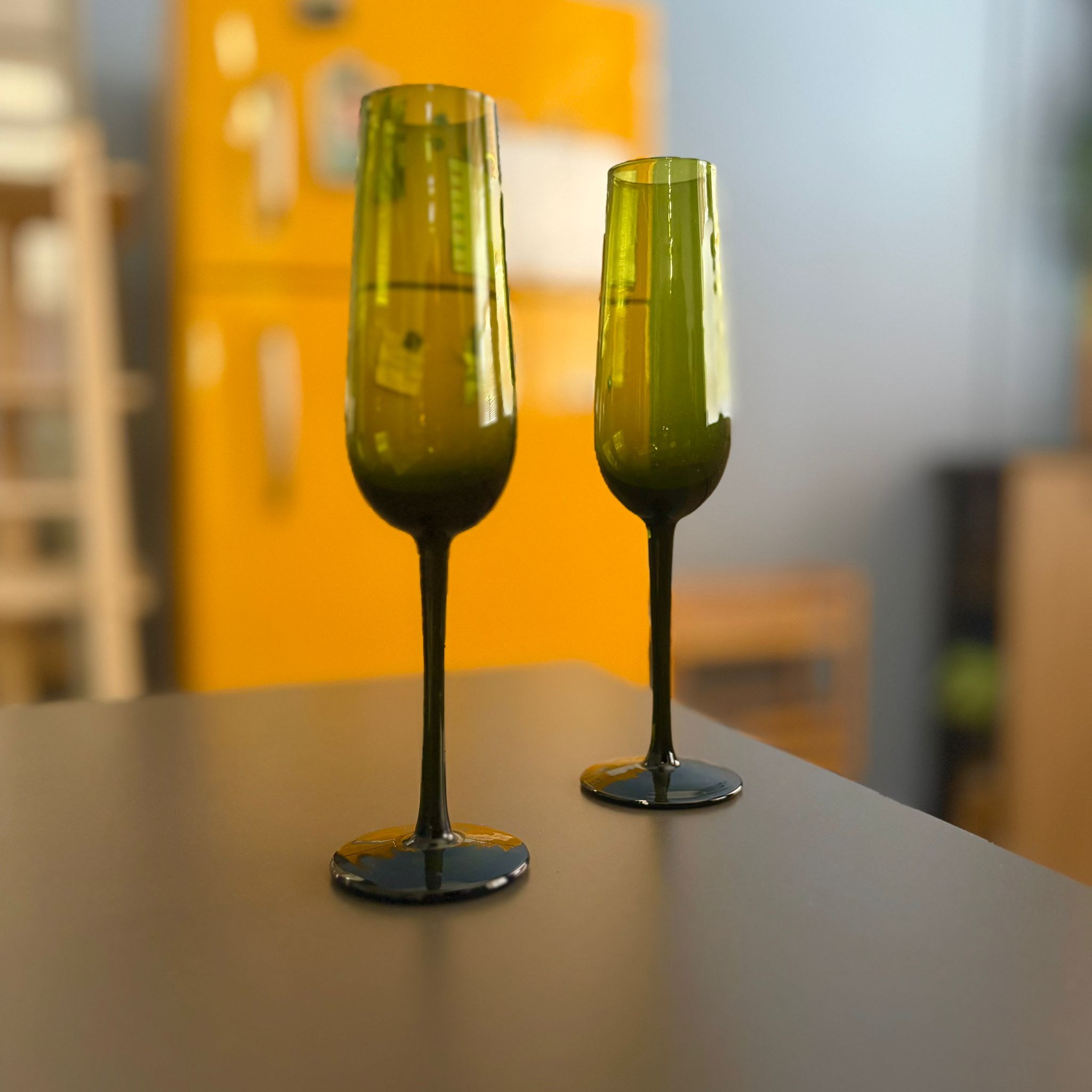 Due calici flute alti in vetro riciclato di colore verde scuro, posizionati su una superficie scura con sfondo giallo sfocato.