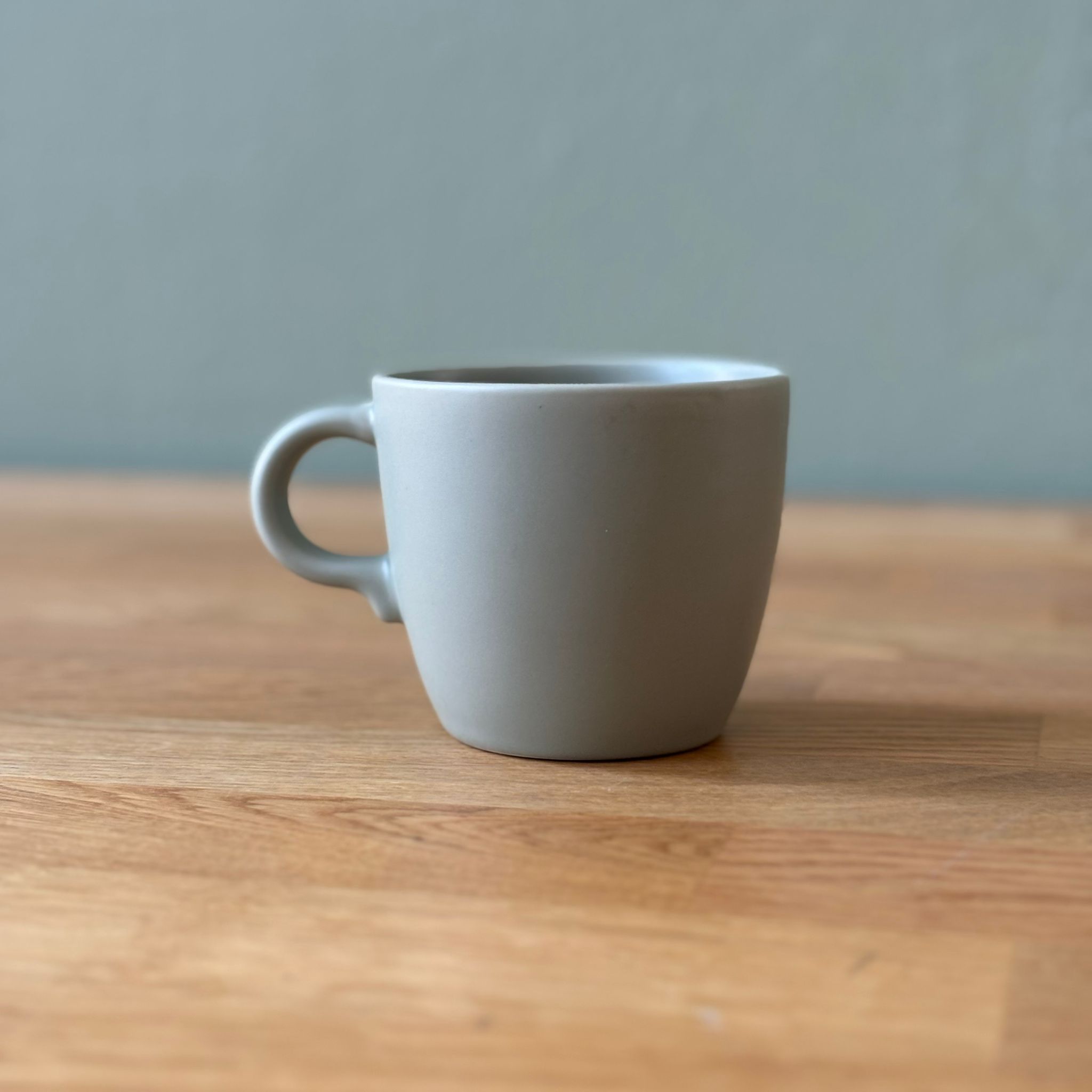 Una tazza in ceramica grigio chiaro opaco con manico, posizionata su una superficie di legno chiaro con sfondo grigio.
