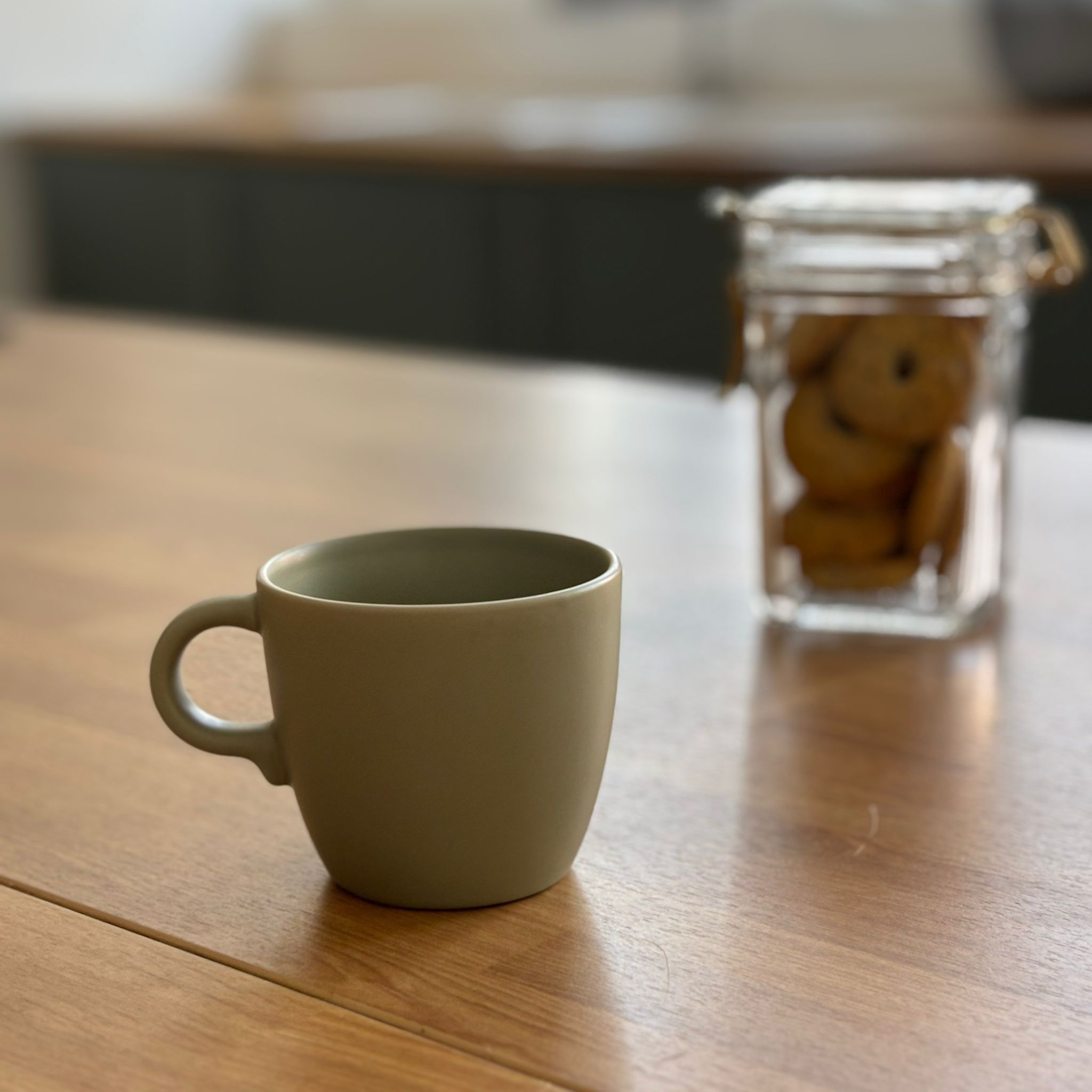 Una tazza in ceramica opaca di colore verde salvia posizionata su un tavolo di legno chiaro, con un barattolo di biscotti sfocato sullo sfondo.