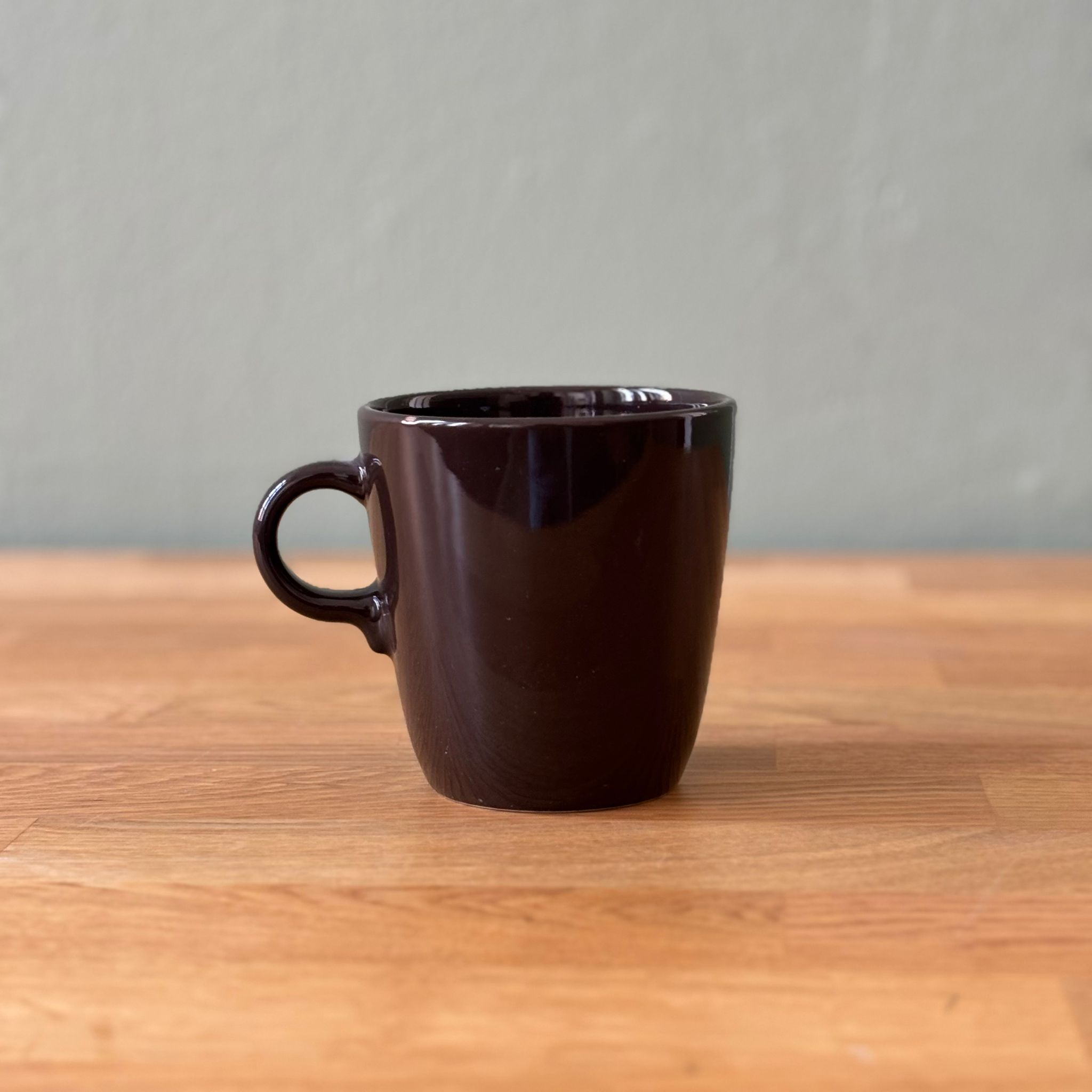 Una tazza in ceramica marrone scuro lucida, con manico, posizionata su un tavolo di legno chiaro con sfondo grigio.