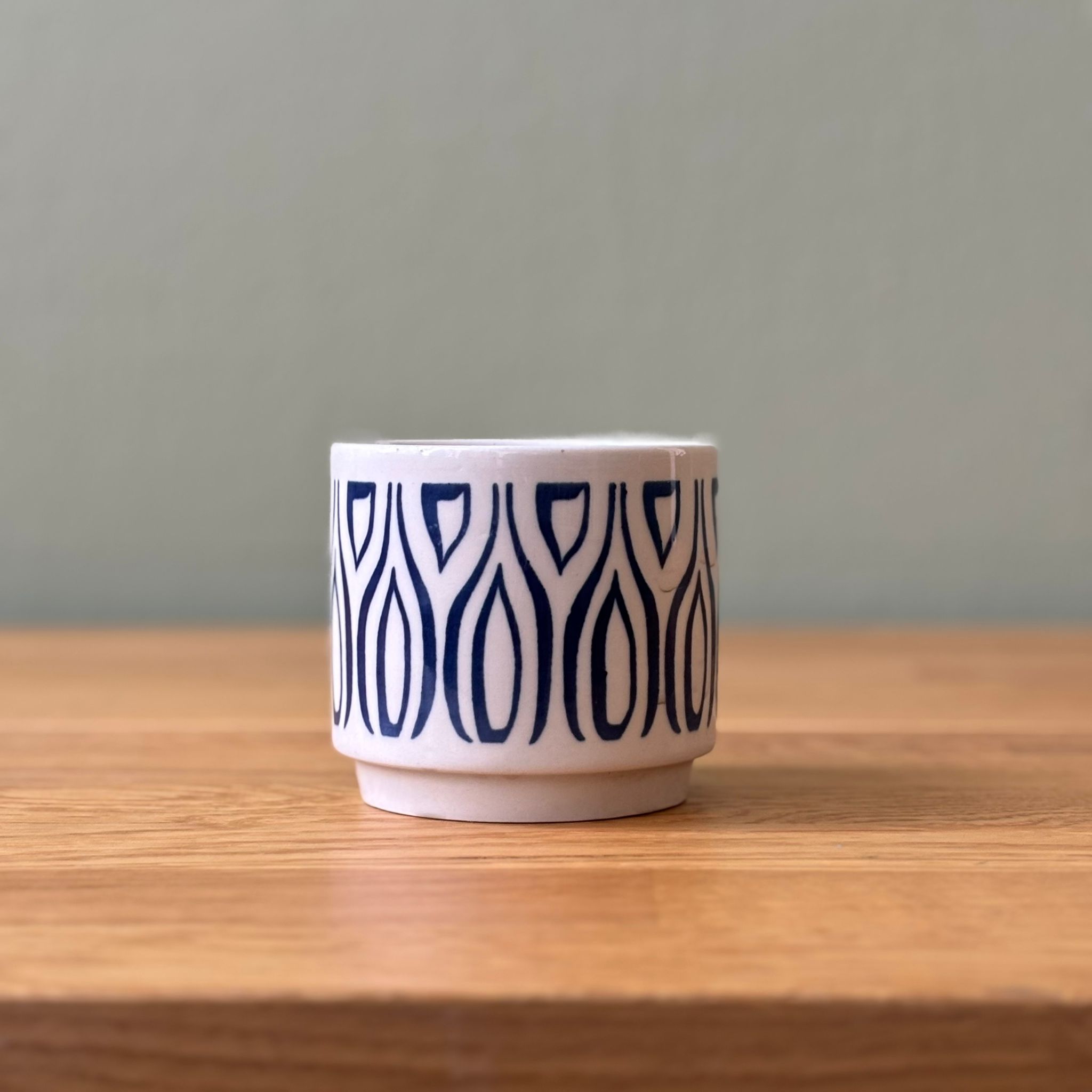 Vaso cilindrico in ceramica bianca con motivo geometrico blu scuro, posizionato su un tavolo di legno chiaro.