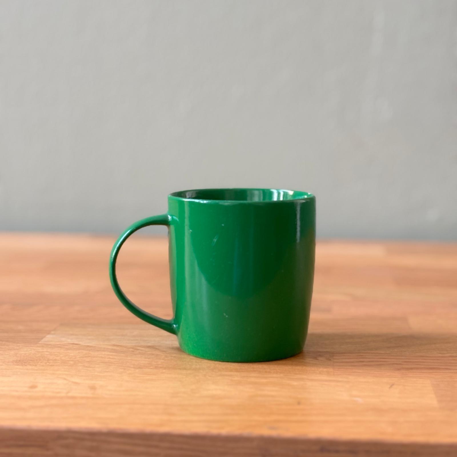 Tazza cilindrica verde brillante con manico, posizionata su un tavolo di legno chiaro con sfondo grigio neutro.