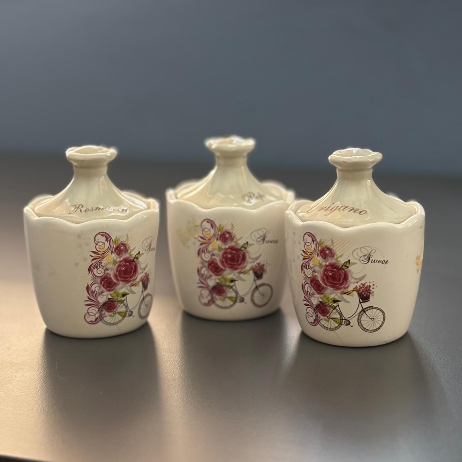 Tre barattoli in ceramica color crema con coperchio, decorati con rose rosse e biciclette, su una superficie scura.