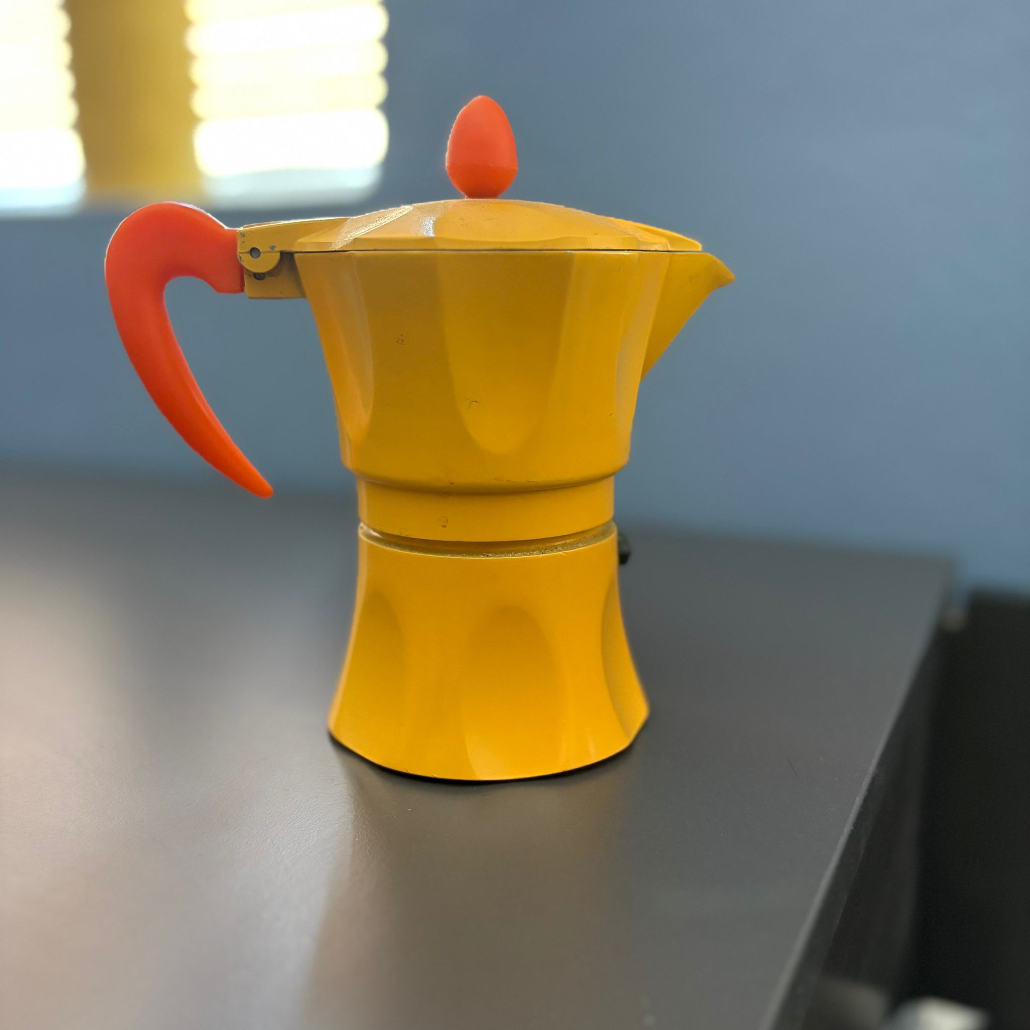Caffettiera Moka di colore giallo brillante con manico e pomello arancioni, posizionata su una superficie scura.