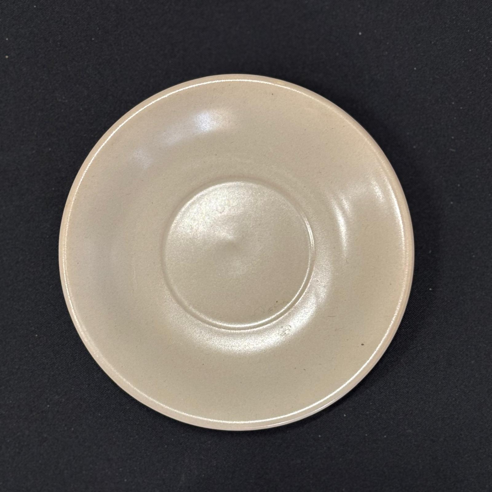 Piattino sottotazza rotondo in ceramica di colore beige chiaro, con bordo leggermente rialzato e incavo centrale, su sfondo scuro.