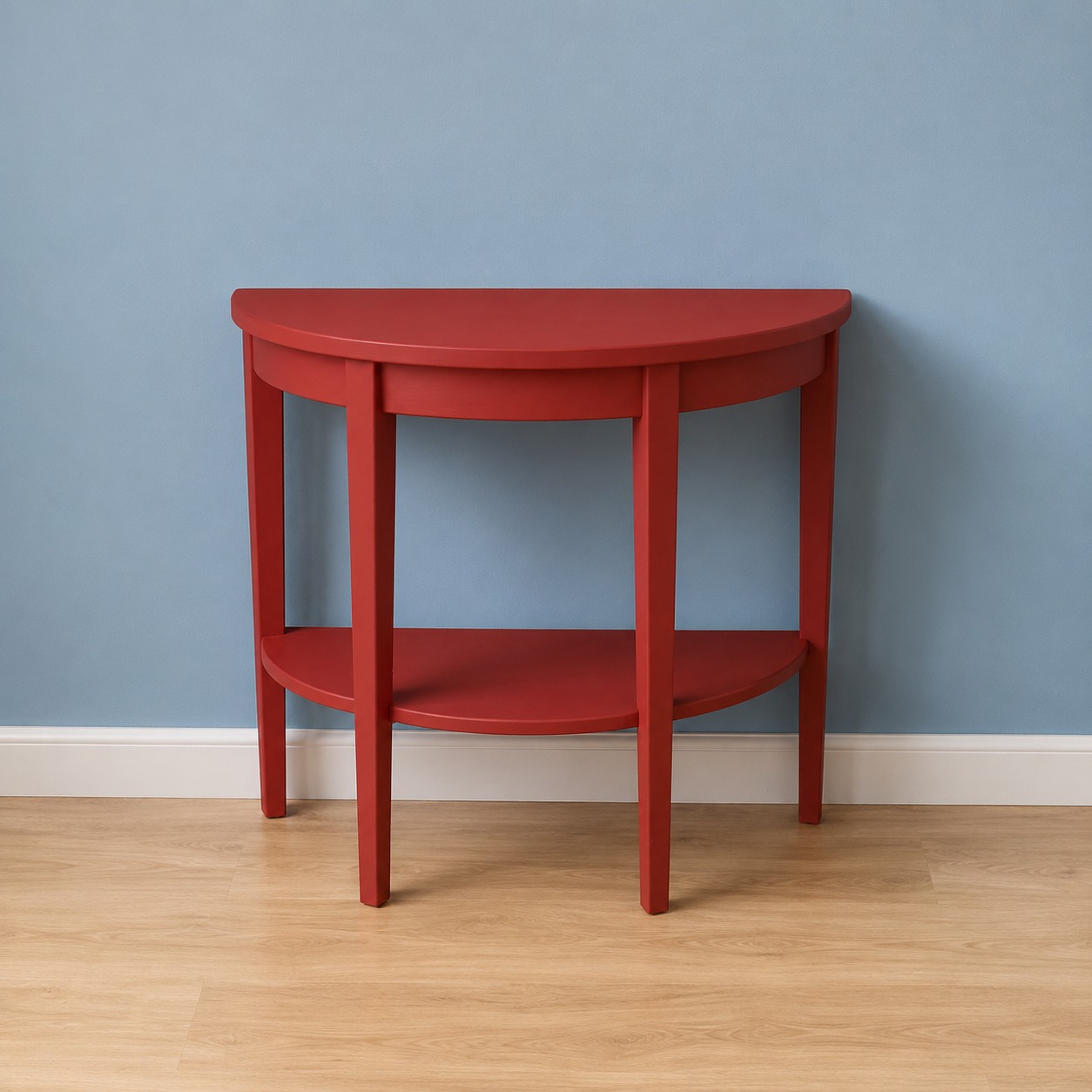 Consolle semicircolare rossa con ripiano inferiore, appoggiata a una parete blu chiaro su pavimento in legno.