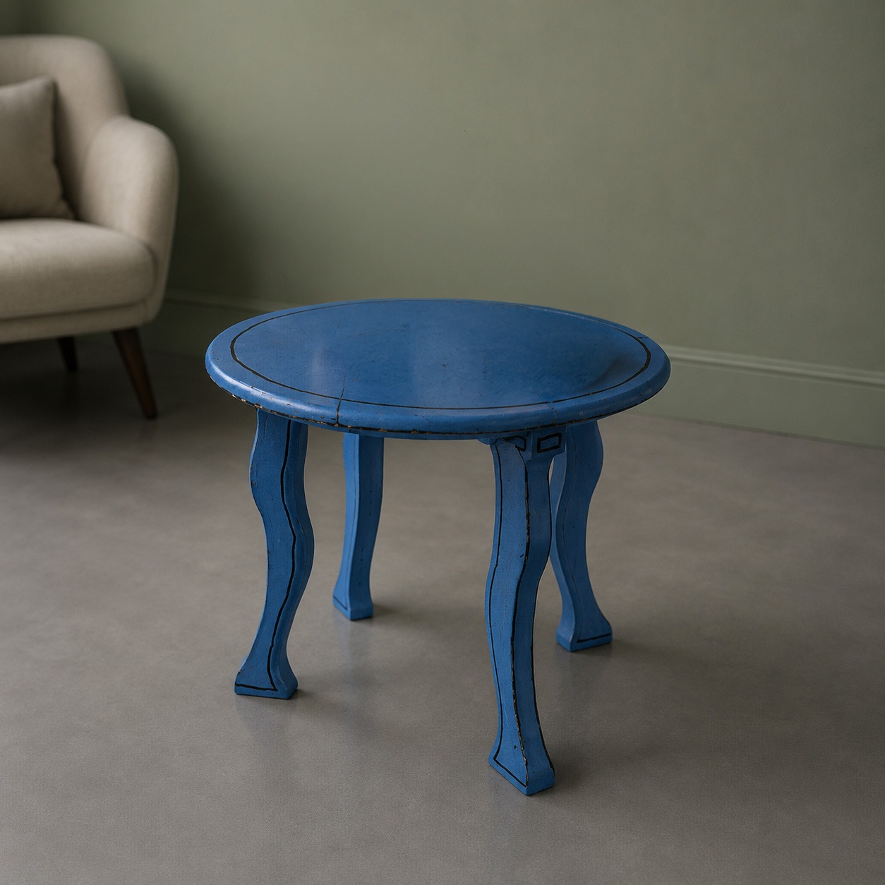 Tavolino rotondo blu con gambe curve e finitura anticata, posizionato su pavimento grigio chiaro accanto a una poltrona beige.