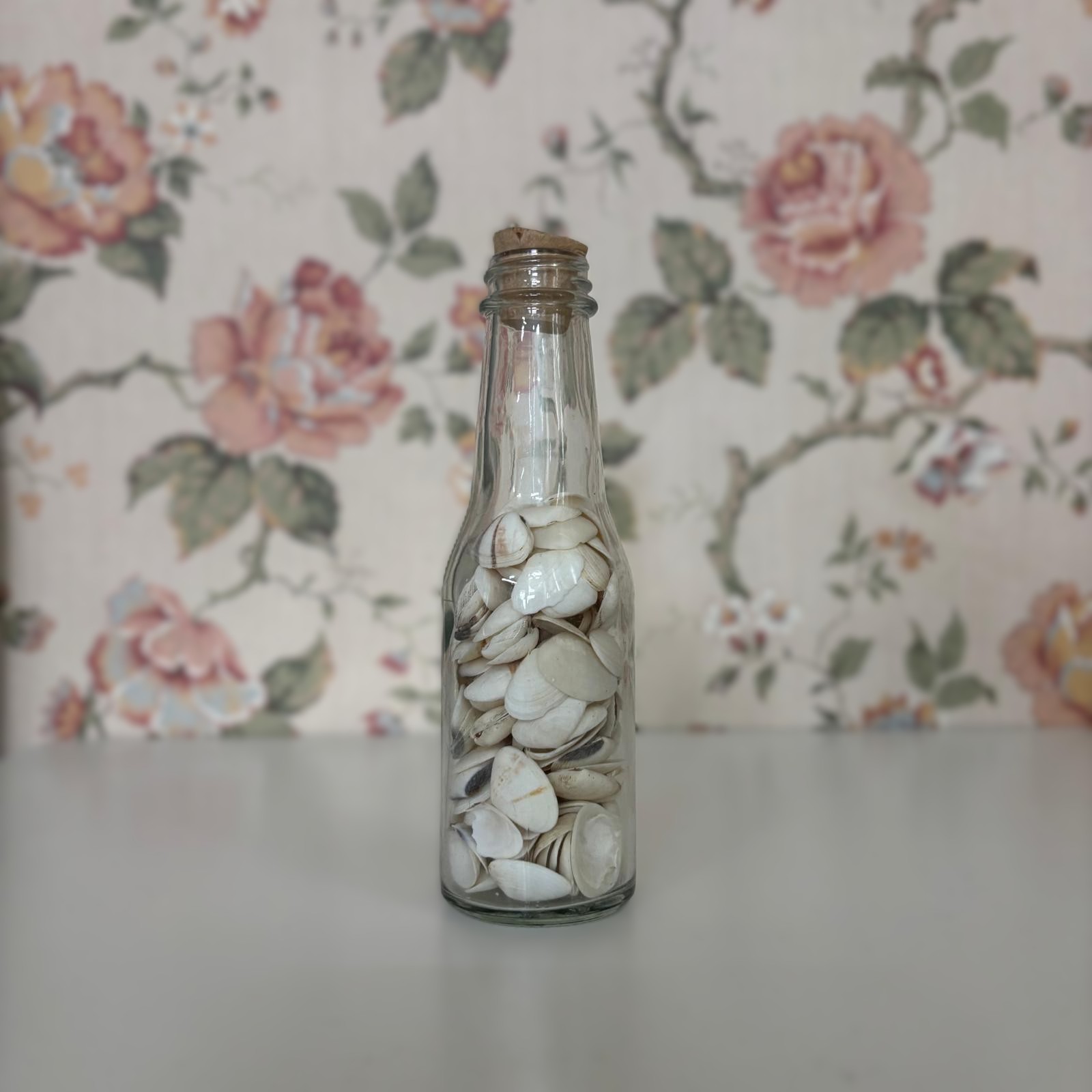 Bottiglia di vetro trasparente riempita di piccole conchiglie bianche e beige, con tappo di sughero, su sfondo floreale.