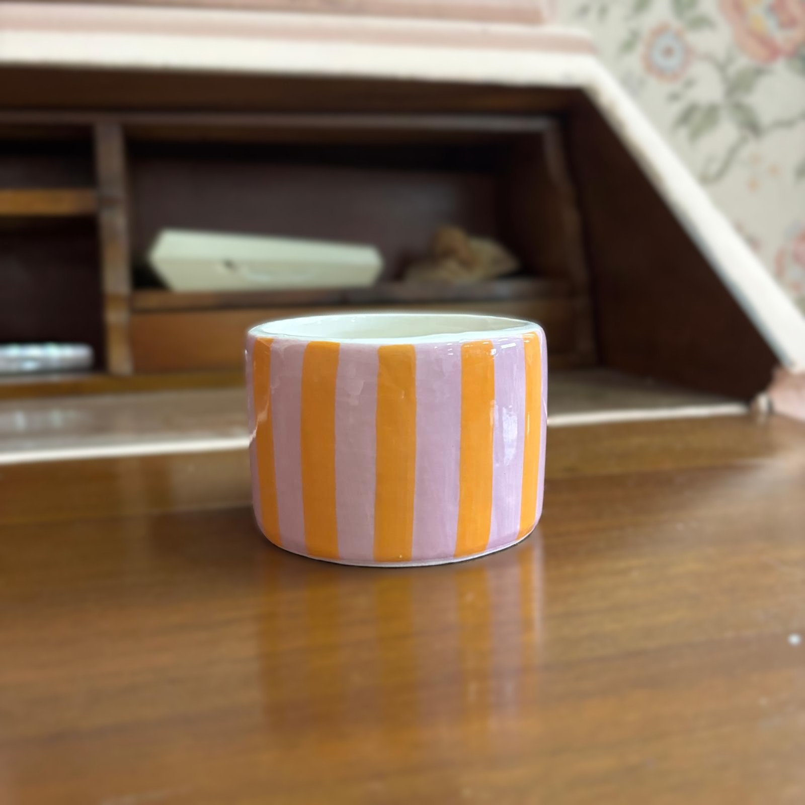 Contenitore cilindrico in ceramica con strisce verticali rosa lavanda e arancioni, posizionato su una superficie di legno.