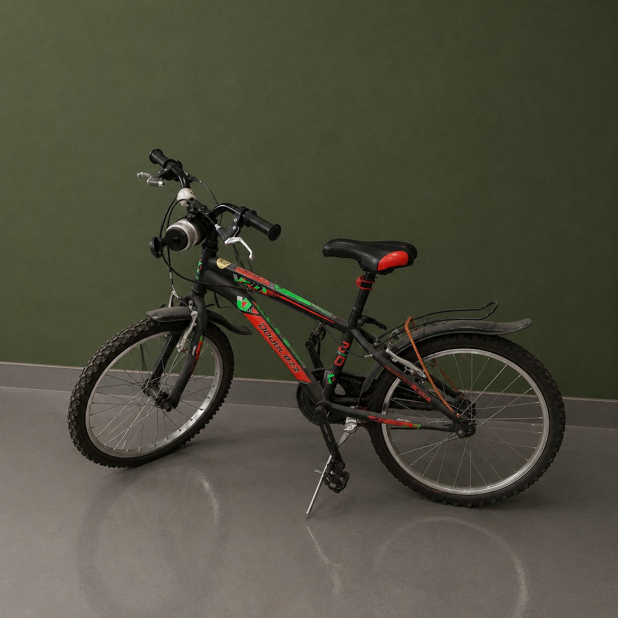 Bicicletta mountain bike nera con dettagli rossi e verdi, parafanghi e portapacchi, appoggiata su cavalletto davanti a parete verde.