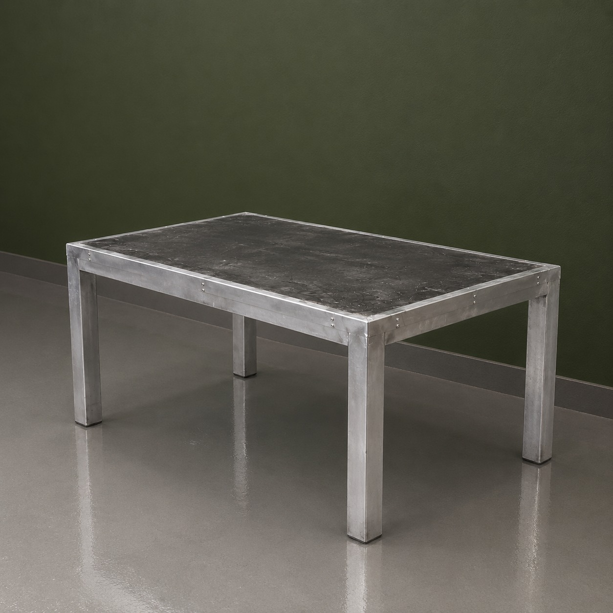 Tavolo rettangolare con struttura in alluminio spazzolato e piano scuro effetto cemento, su pavimento grigio con parete verde scuro.