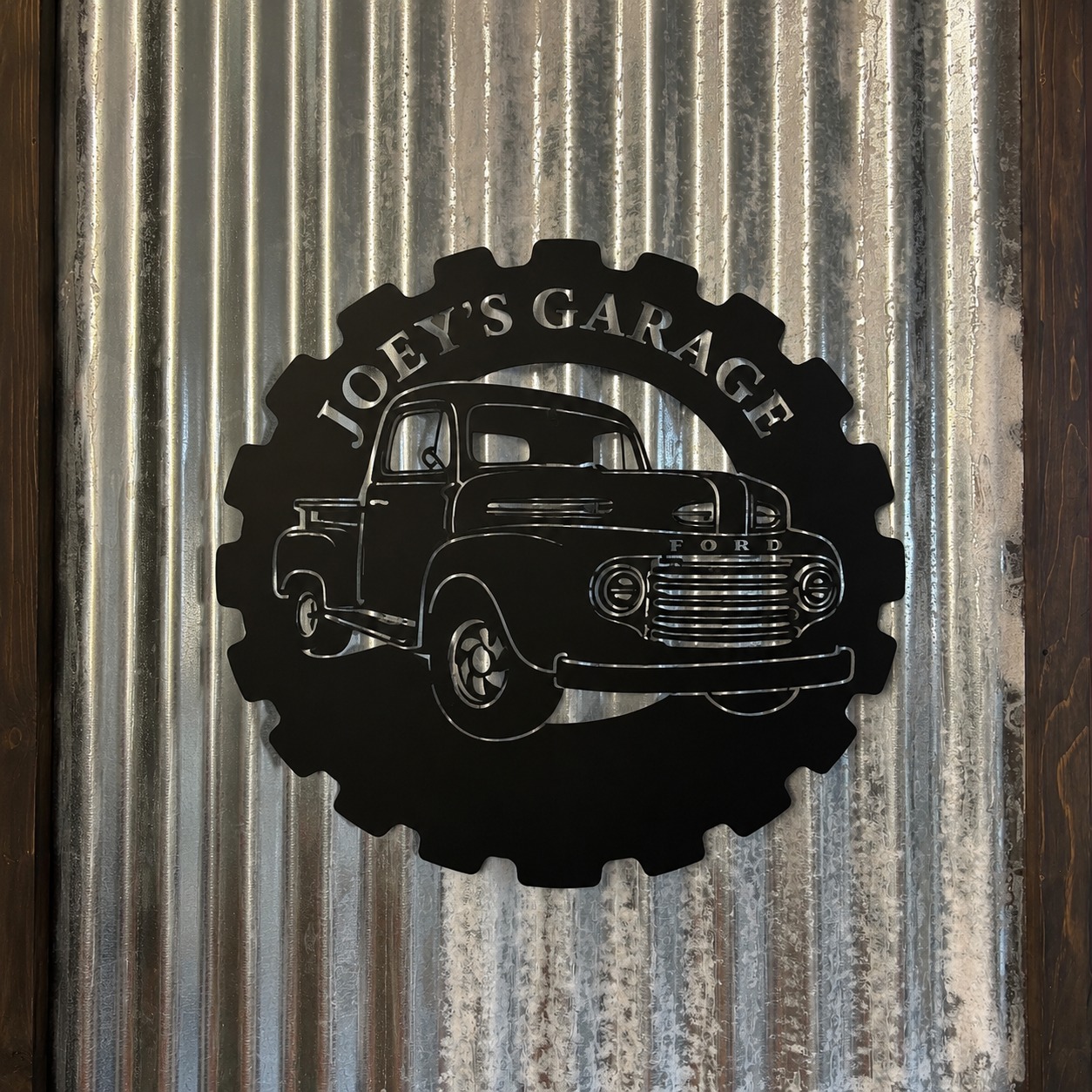 Insegna decorativa circolare in metallo nero a forma di ingranaggio con silhouette di pickup Ford e scritta "Joey's Garage", montata su lamiera ondulata.