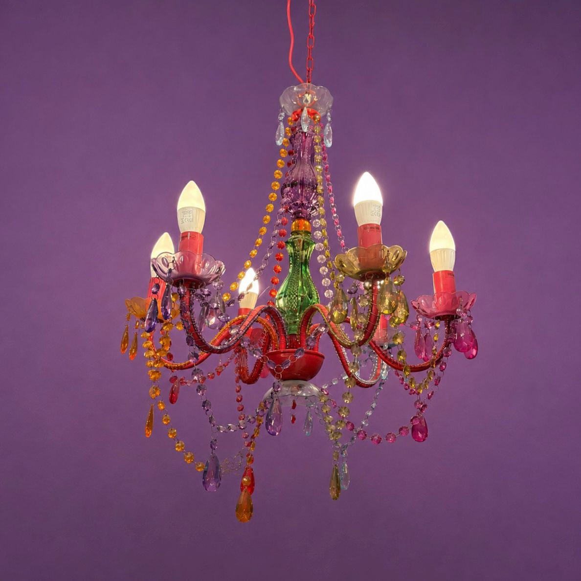 Lampadario eclettico a sei bracci con struttura rossa, decorato con perline e pendenti in vetro e acrilico multicolore su sfondo viola.