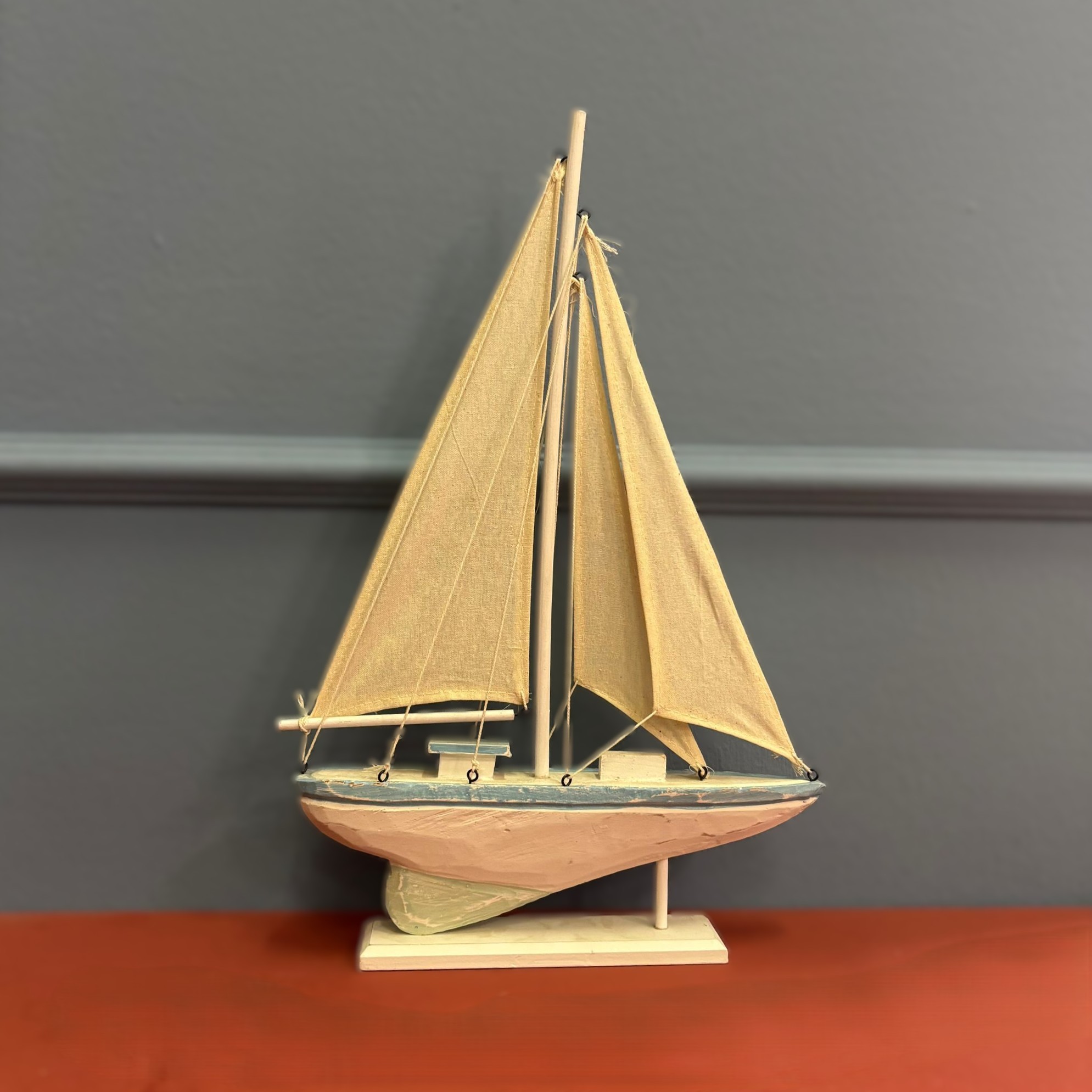Modello di veliero decorativo in legno con scafo bianco e azzurro, vele beige, su base bianca, contro parete grigia.