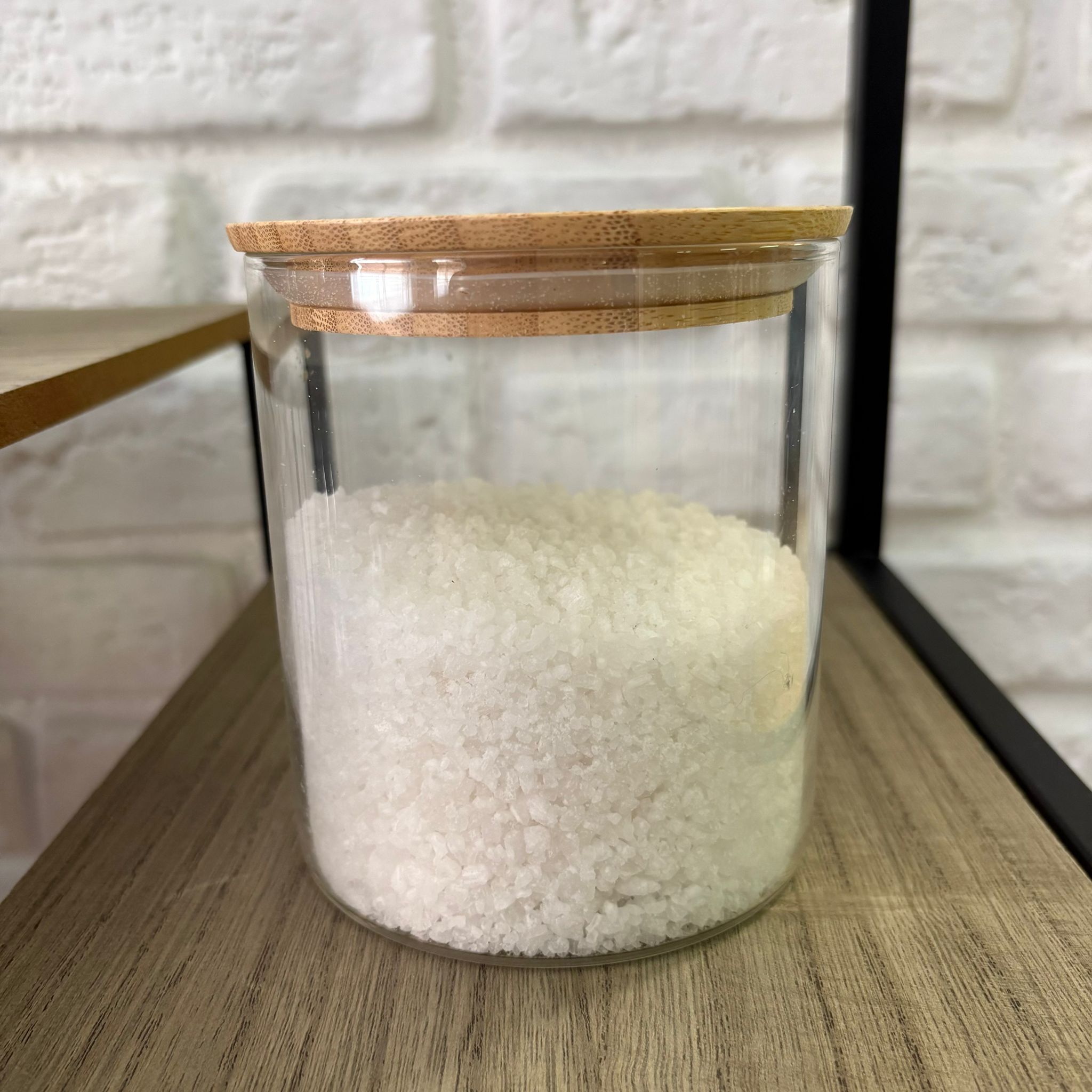 Barattolo cilindrico in vetro trasparente con coperchio in bambù, riempito di sale grosso bianco, posizionato su una mensola in legno.