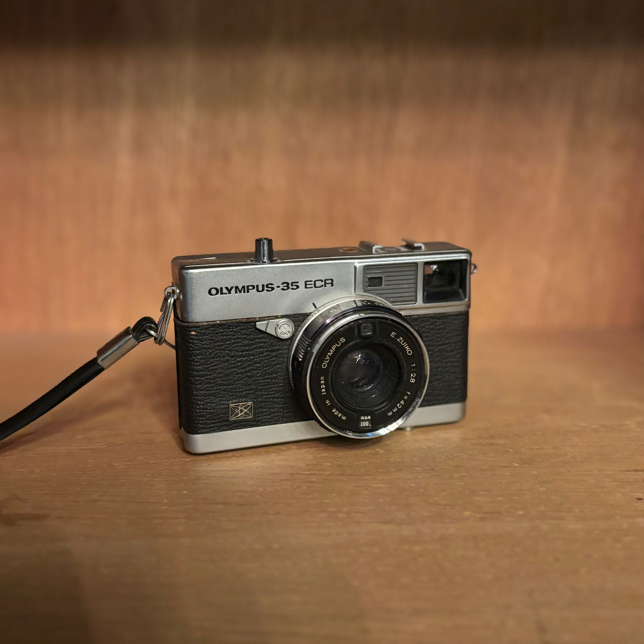 Fotocamera vintage Olympus-35 ECR con corpo cromato e rivestimento nero, obiettivo E. ZUIKO, su superficie di legno.